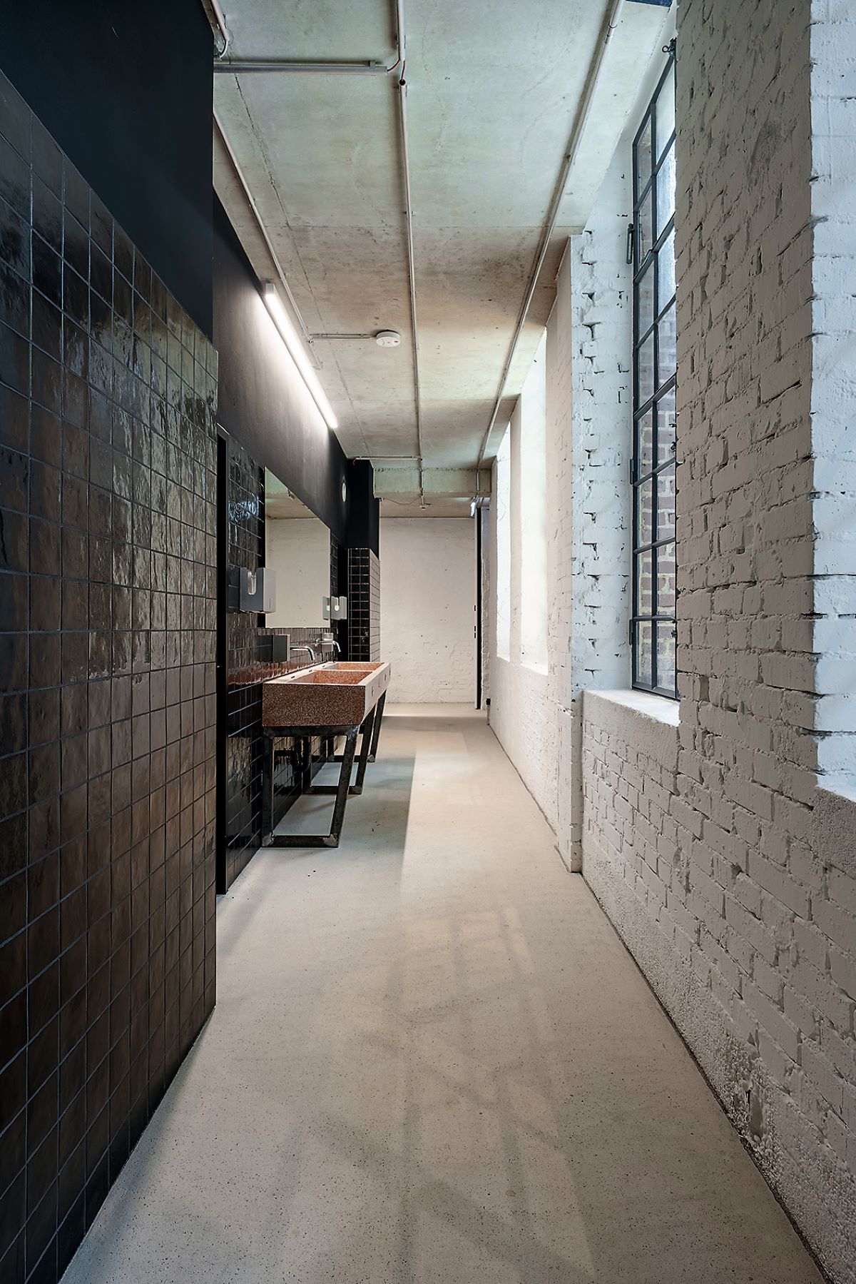 Sanitärbereich mit einem schalen Flur in modernem Industriestil mit weiß gestrichenem Ziegelmauerwerk rechts und einer schwarzen, gefliesten Wand links. Auf der linken Seite steht ein Waschtisch aus Terazzo-Material. Große Fenster rechts lassen Tageslicht in den Raum. Die Decke besteht aus Sichtbeton mit sichtbaren Leitungen.
