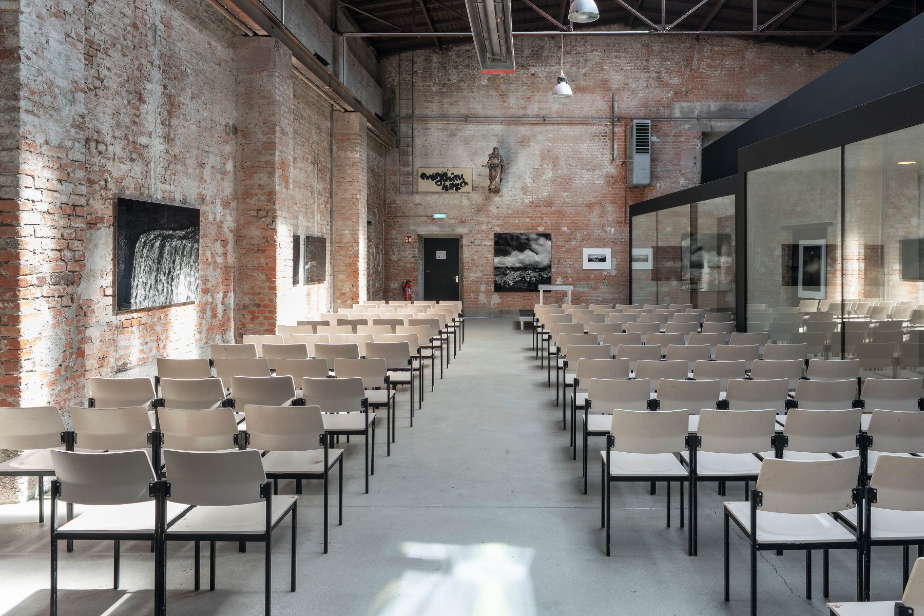 Industriehalle mit rohen Backsteinwänden, eingerichtet als Veranstaltungsraum. Reihen heller Stühle füllen den Raum, ausgerichtet auf eine schwarze Tür im Hintergrund. Rechts verläuft eine Glaswand. Oben sind sichtbare Rohre und Beleuchtungselemente.