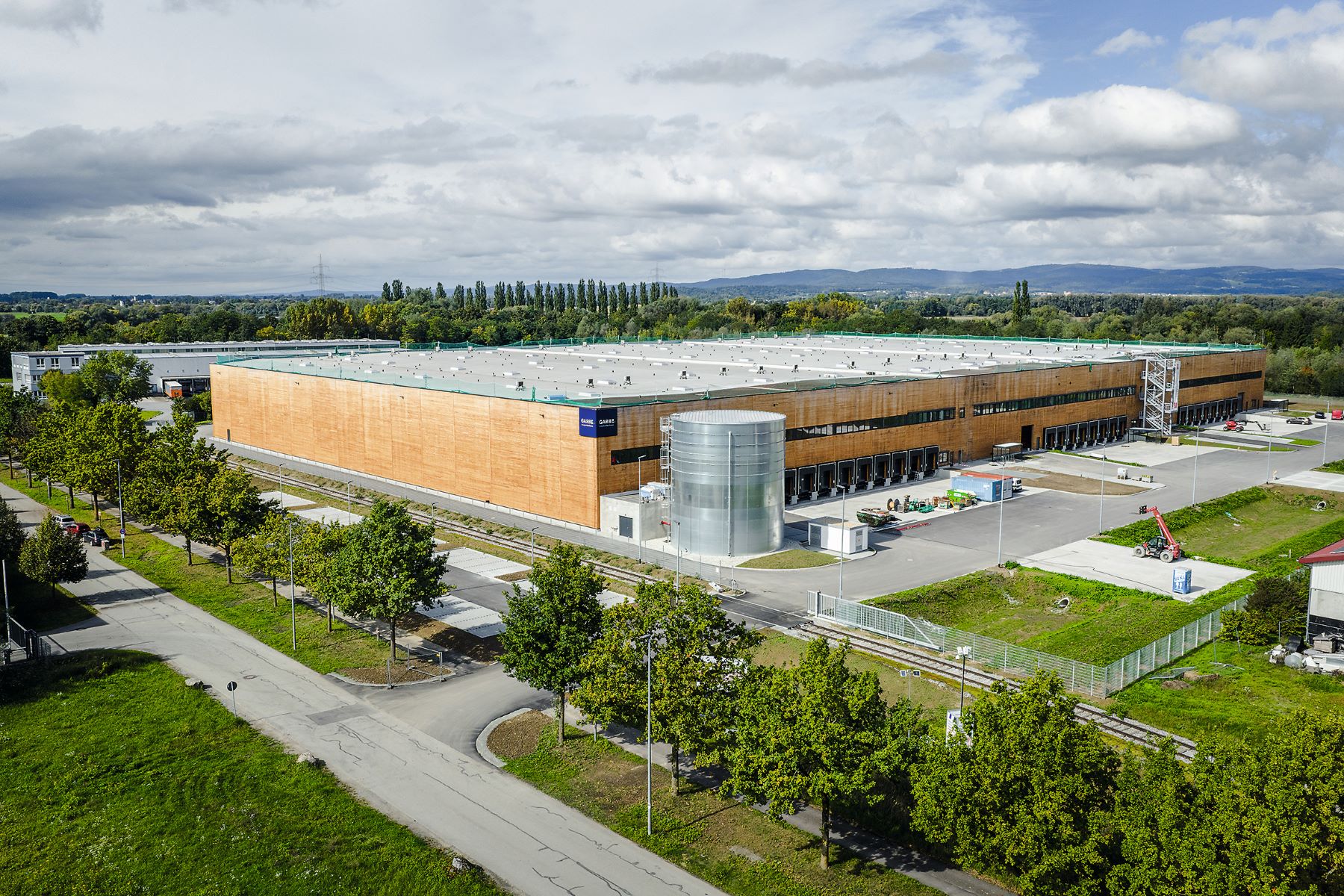 Luftaufnahme einer großen Logistikhalle mit Holzfassade und Flachdach. Vor der Halle befinden sich breite Zufahrts- und Ladebereiche, daneben Grünflächen und Bäume. Im Hintergrund leicht bewaldete Hügel und teils bewölkter Himmel.