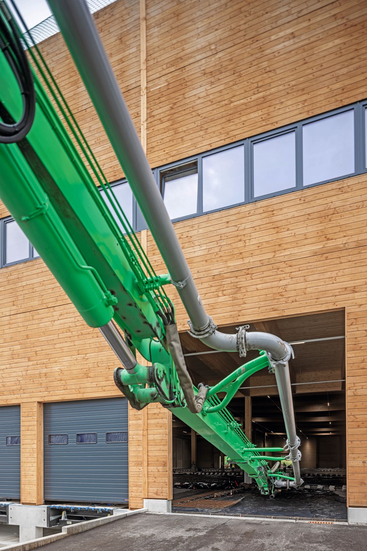 Grüner Betonpumpausleger führt durch ein Tor in eine Halle. Im Vordergrund ist eine Holzfassade mit Fenstern zu sehen, darunter mehrere geschlossene Ladetore. Die Betonpumpe wird zum Einbringen von Beton im Innenbereich genutzt. 