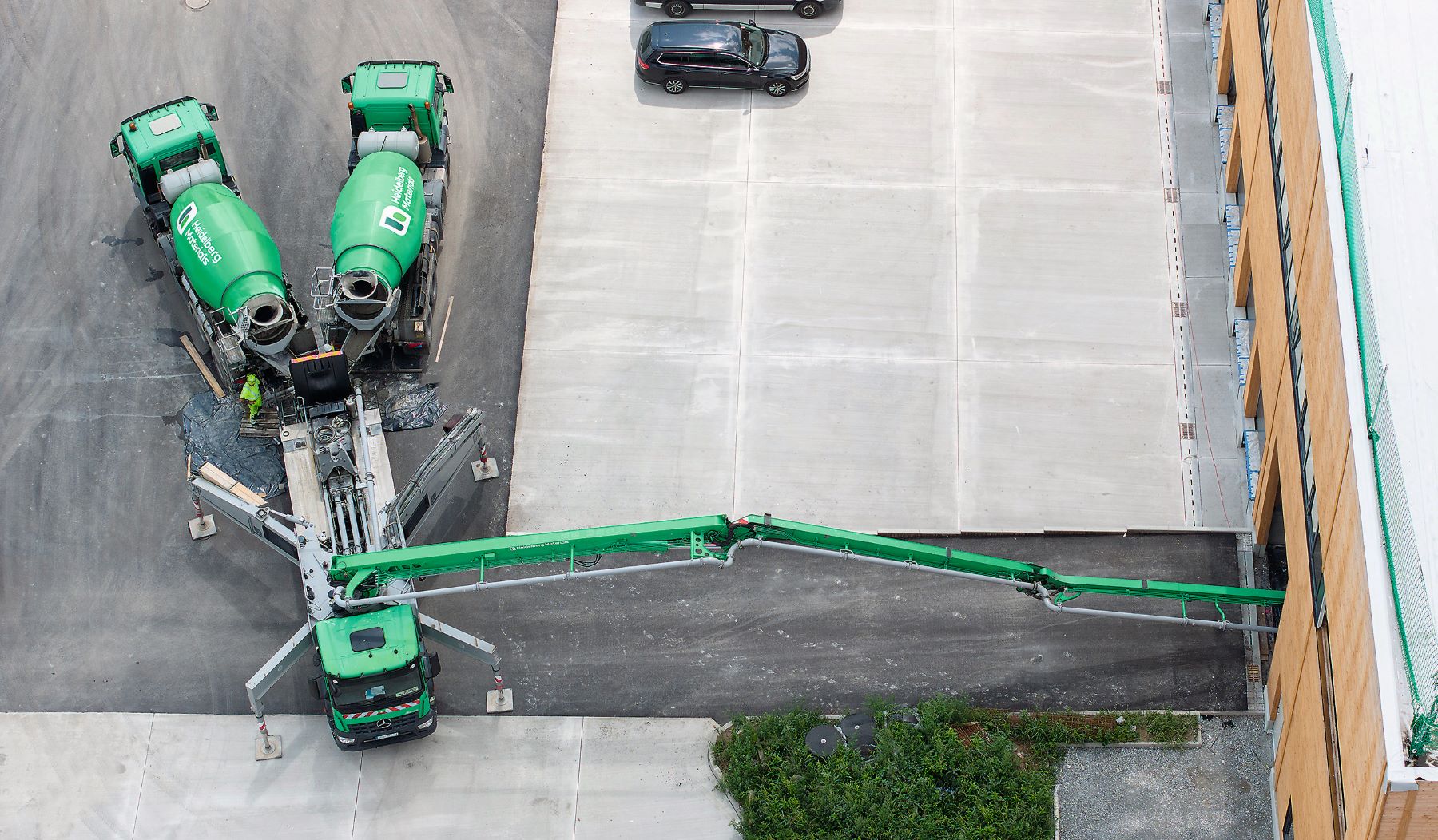 Drohnenaufnahme mehrerer grüner Fahrmischer und eines Pumpenfahrzeugs von Heidelberg Materials. Der Pumpenausleger führt in das Gebäude, daneben befinden sich asphaltierte und begrünte Flächen.