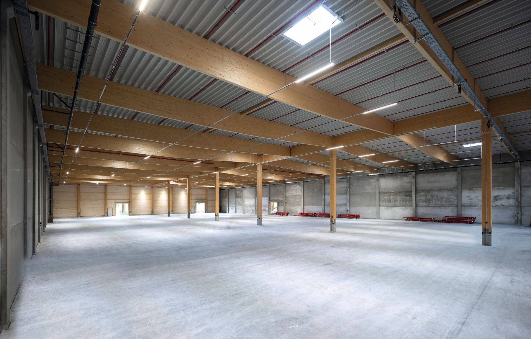 Große, leere Lagerhalle mit Holzstützen, Sichtbetonwänden und einer Stahlträgern und Lichtöffnungen. Der Boden ist betoniert und unbelegt; auf der rechten Seite liegen rote Bauelemente. Im Hintergrund befinden sich mehrere Tore. Tageslicht fällt durch Öffnungen im Dach ein.