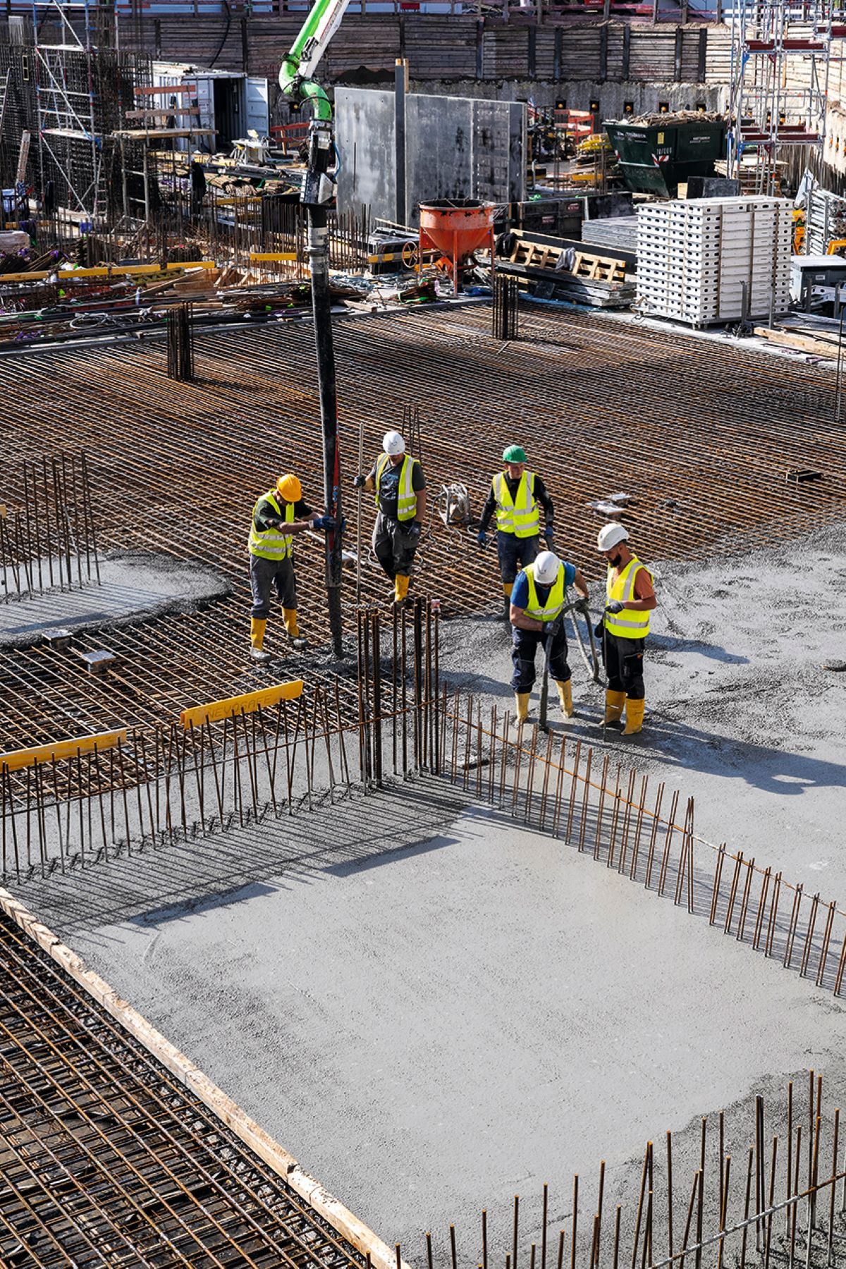 Baustelle mit Arbeitern in Schutzkleidung und Helmen, die Beton auf eine vorbereitete Fläche gießen. Im Vordergrund sind Stahlarmierungen sichtbar. Im Hintergrund Baugerüste, Materialien und Maschinen.