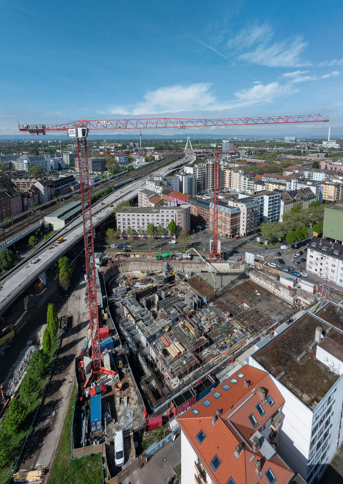 Luftaufnahme einer großen Baustelle mit einem roten Turmdrehkran in der Mitte. Umgeben von Wohn- und Geschäftsgebäuden, Staßen sowie einer erhöhten Verkehrsbrücke auf der linken Seite. Im Vordergrund Baustellenarbeiten und Baumaterialien, im Hintergrund grüne Flächen und ein blauer Himmel mit wenigen Wolken.