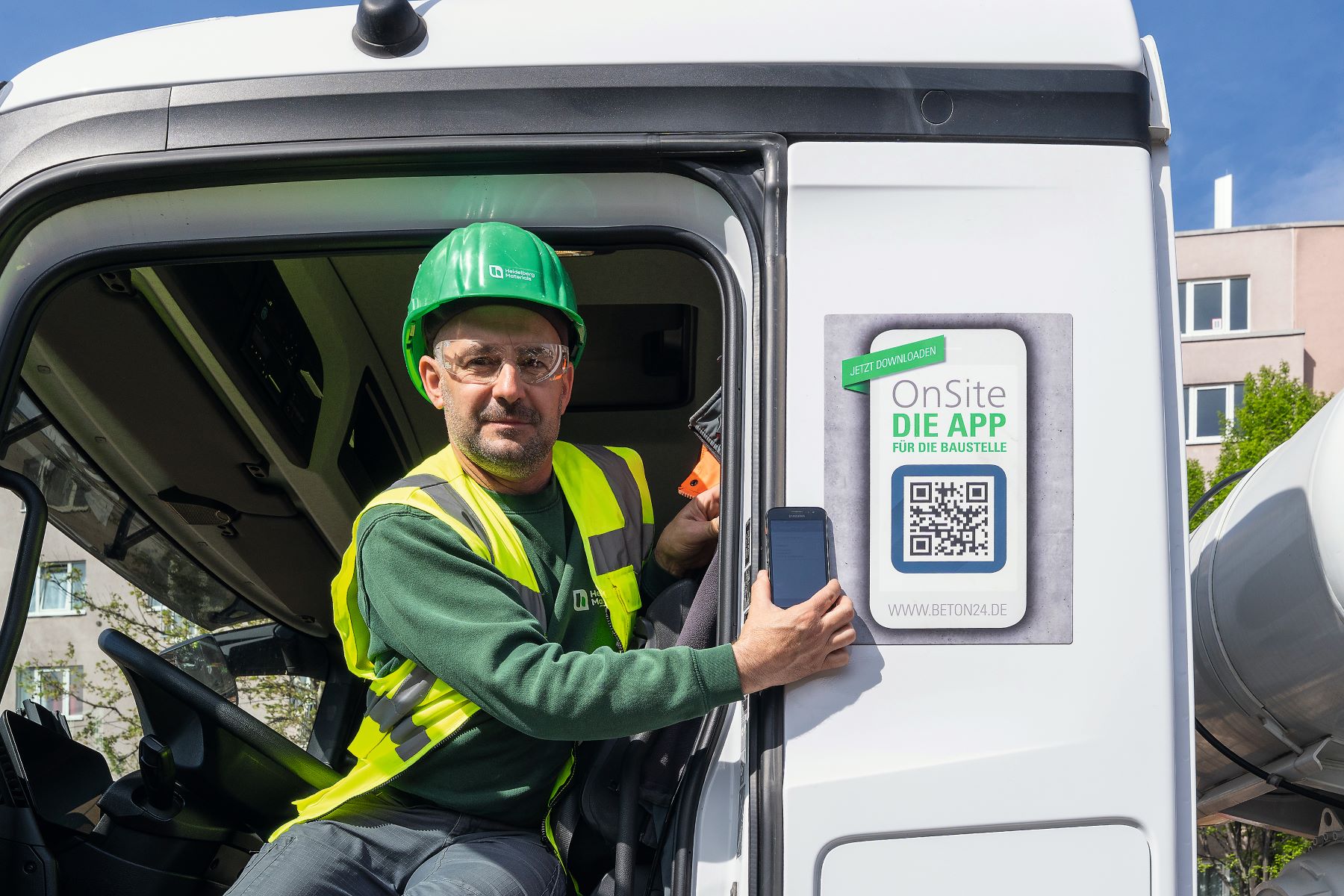 Ein Bauarbeiter mit grünem Helm und Warnweste sitzt in der offenen Fahrerkabine eines Fahrzeugs und hält ein Smartphone, auf dem die Onsite-App gezeigt wird. Außen am Kabinenrahmen befindet sich ein Aufkleber mit QR-Code und der Aufschrift "Onsite - Die App für die Baustelle".