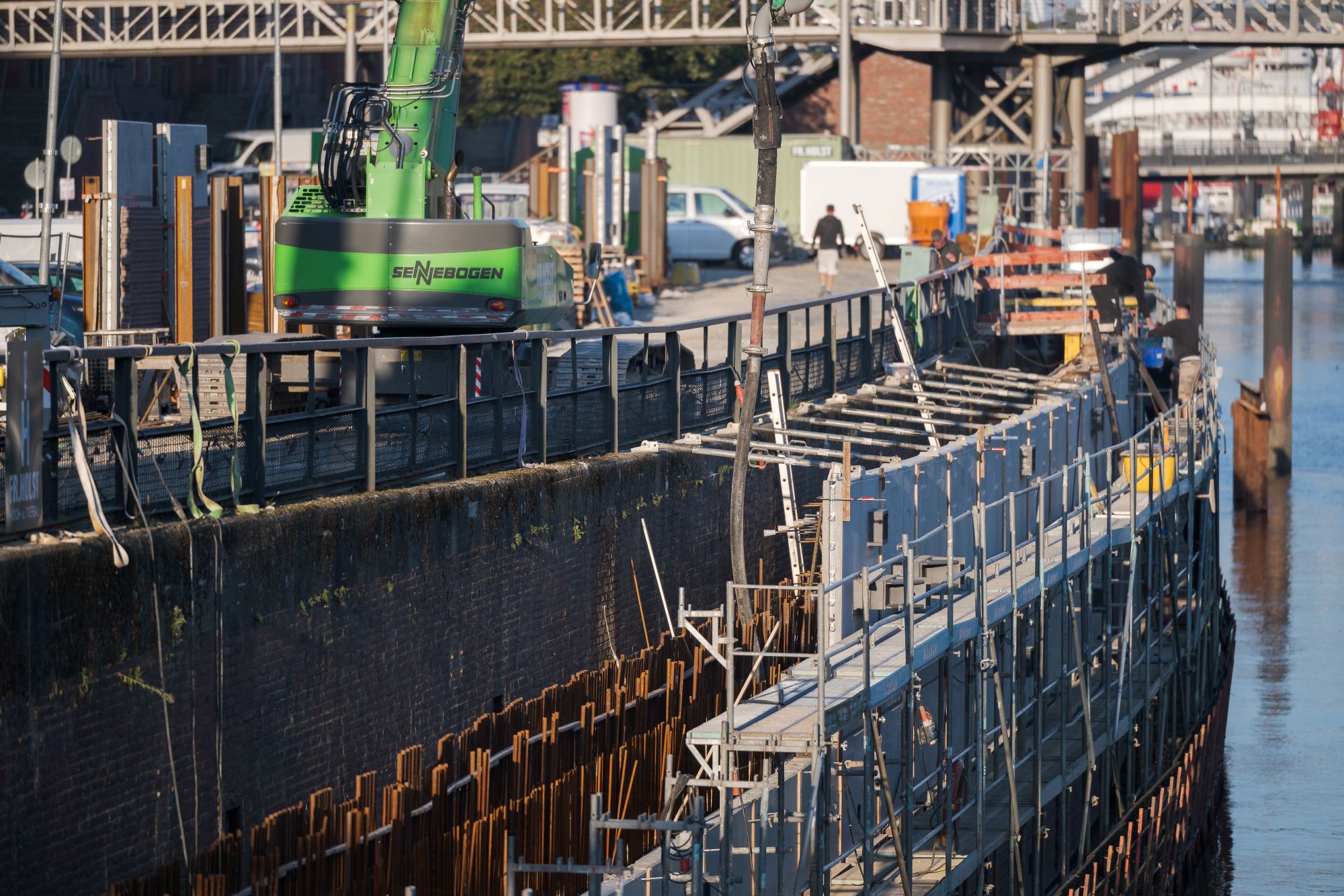 Bauarbeiten an einer Uferbefestigung mit Gerüsten und einem grünen Bagger im Einsatz.