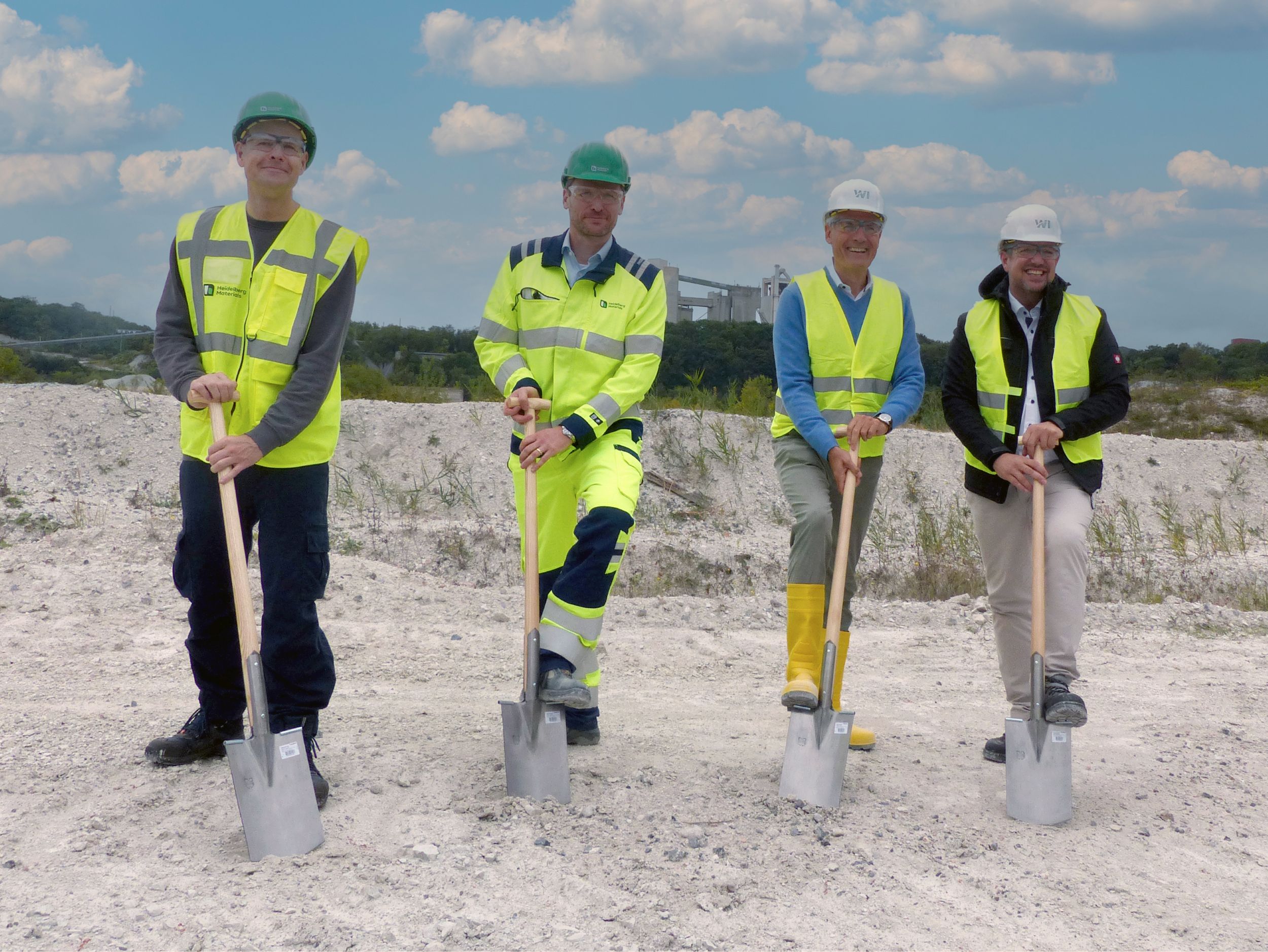Vier Personen mit Schutzkleidung halten Spaten auf einer Baustelle.