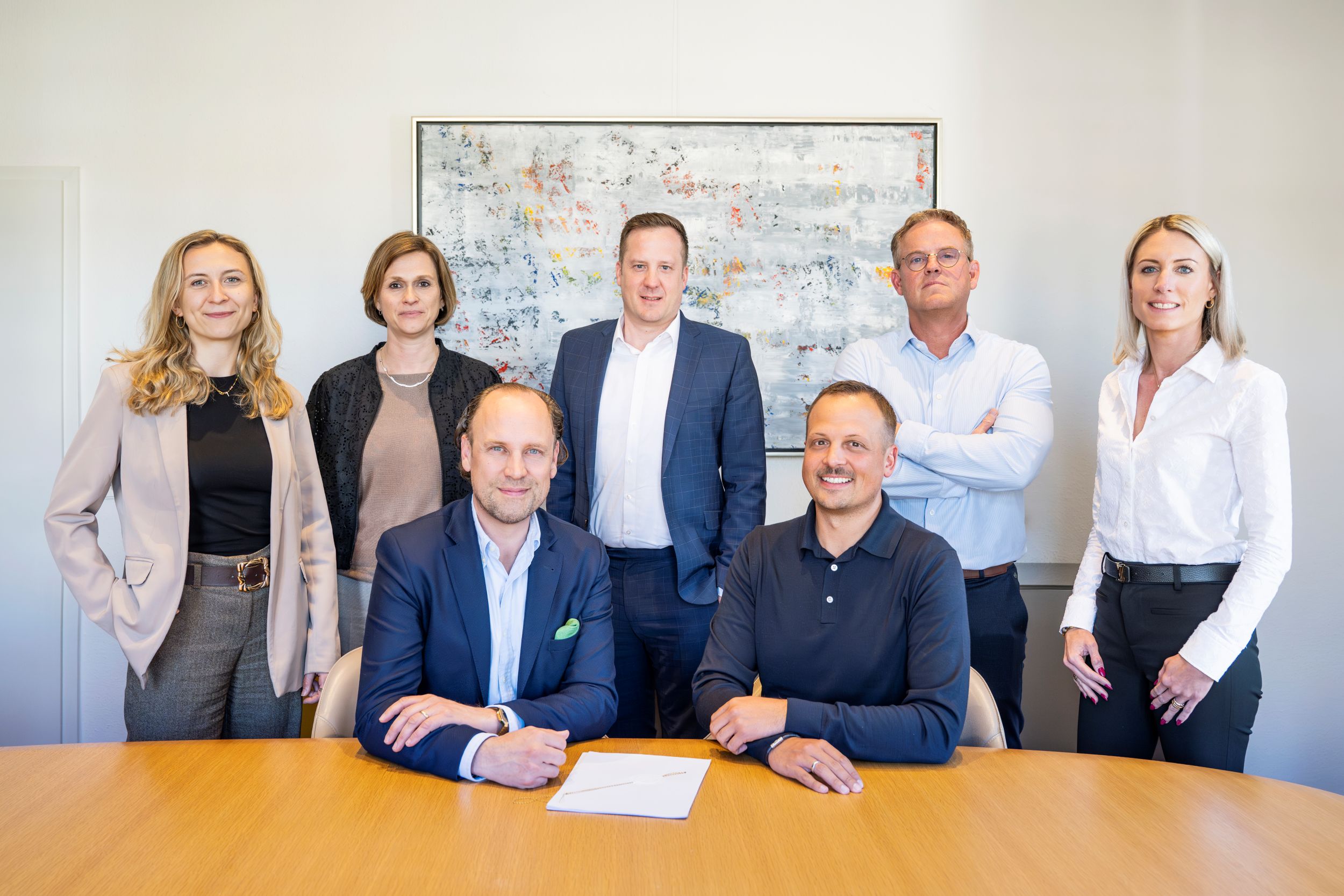 Mehrere Personen in Businesskleidung stehen und sitzen an einem Konferenztisch in einem Büro.
