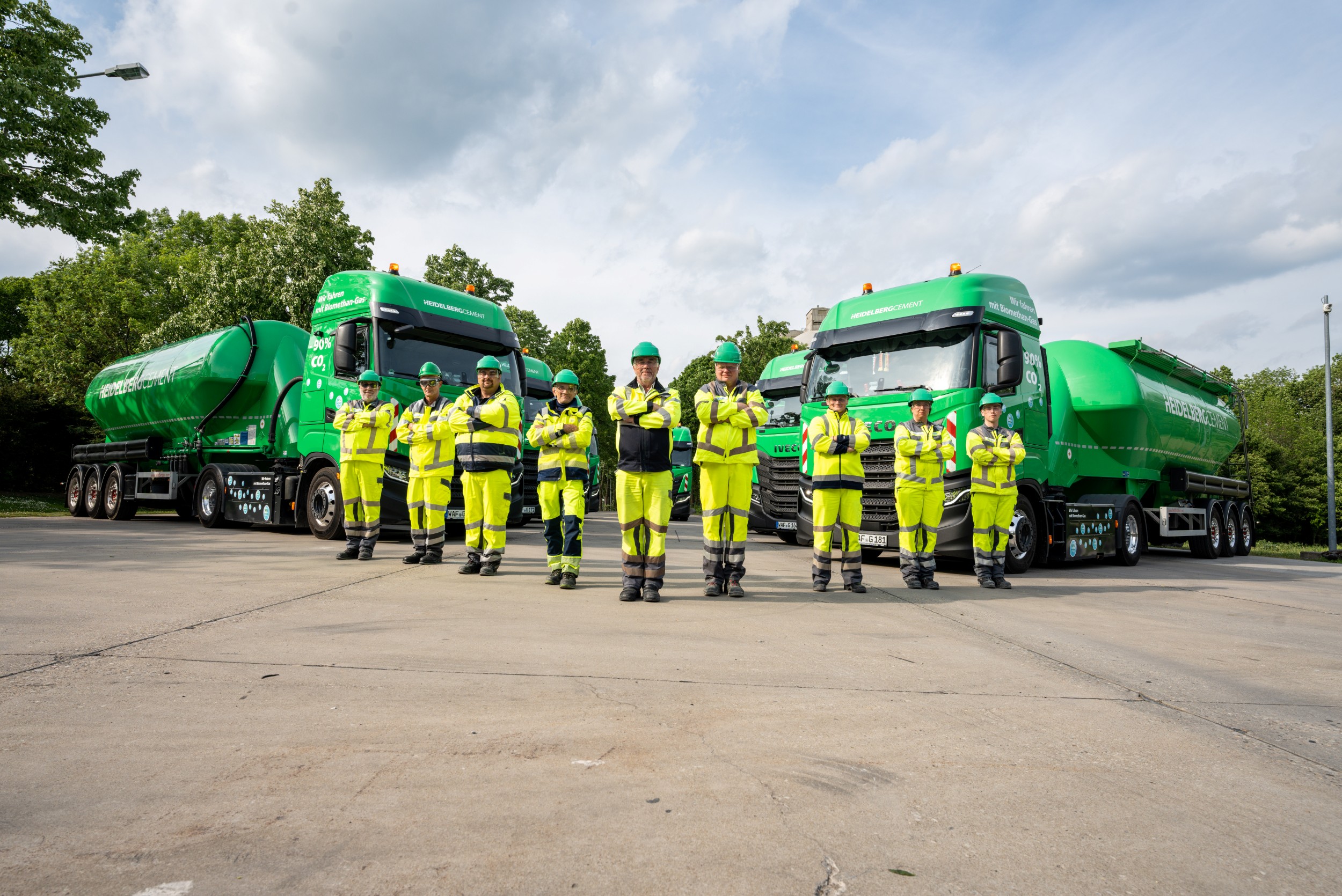 Gruppe von neun Personen in gelber Arbeitskleidung und Schutzhelmen vor zwei grünen Silozügen. Im Hintergrund sind Bäume und ein bewölkter Himmel sichtbar.