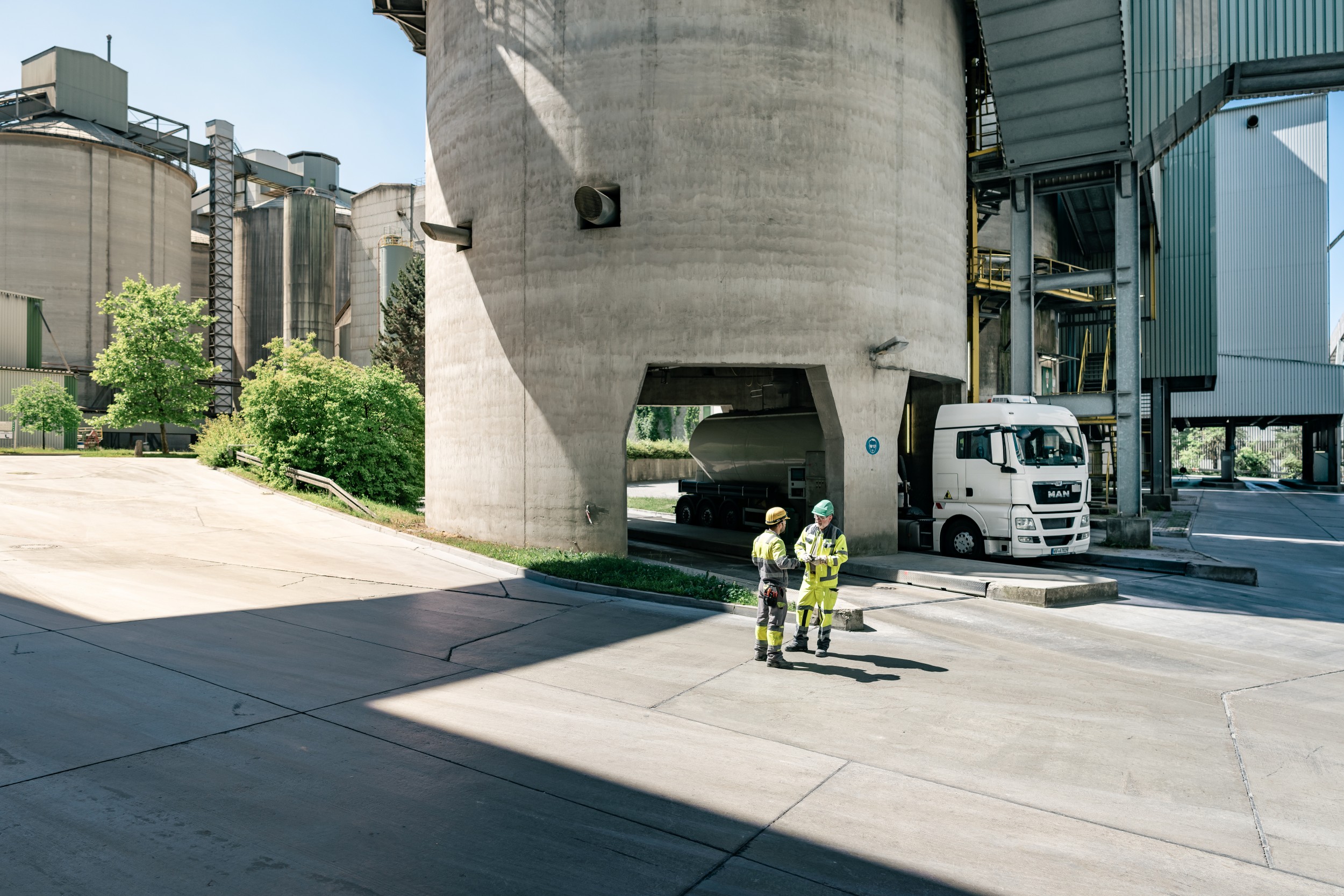 Zwei Arbeiter in Schutzkleidung stehen auf einem Zementwerk und unterhalten sich; im Hintergrund ein weißer Silozug.