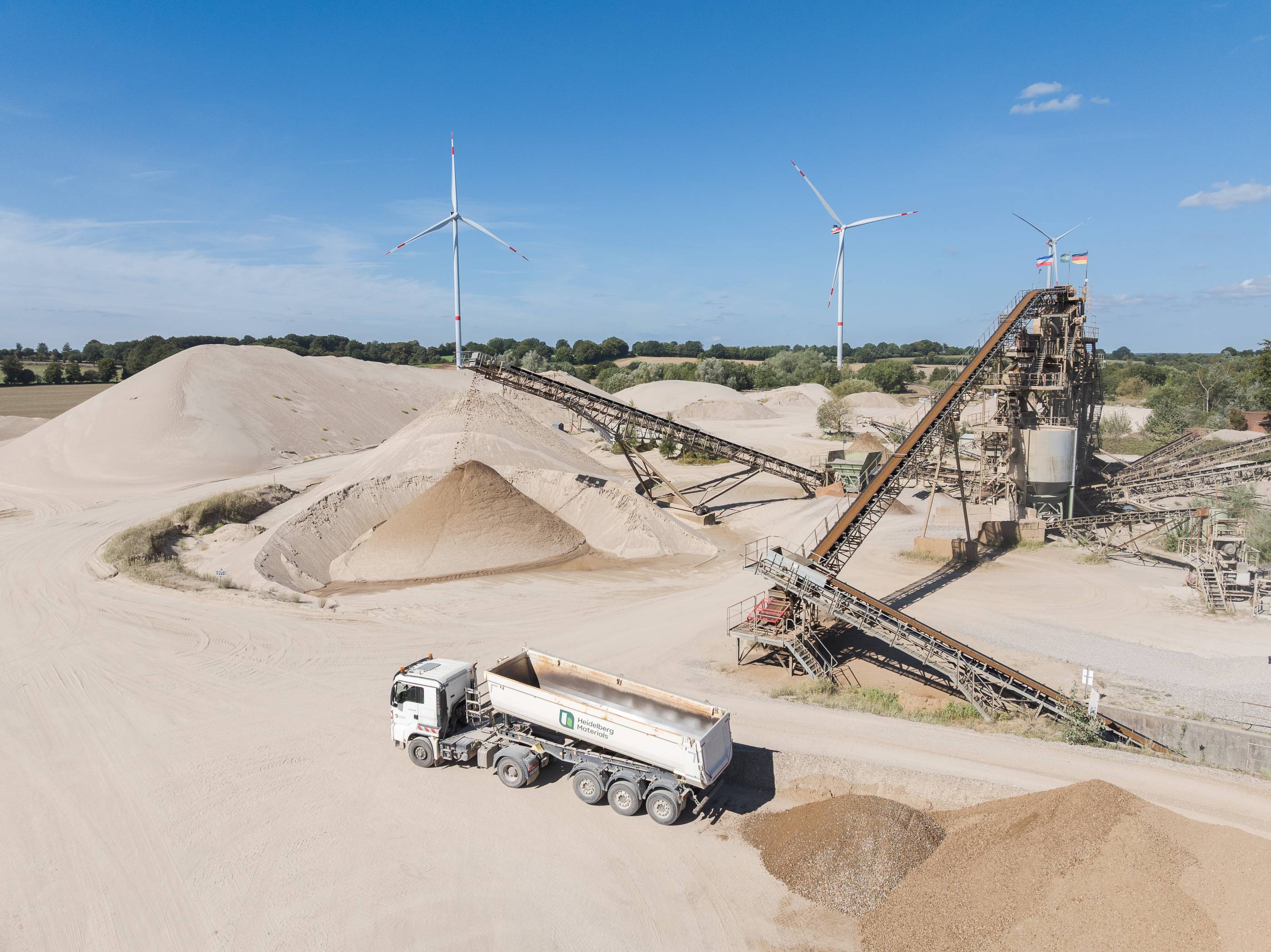 Ein Industriegelände mit Kieshaufen, Förderbändern und einem Lastwagen, im Hintergrund Windräder.