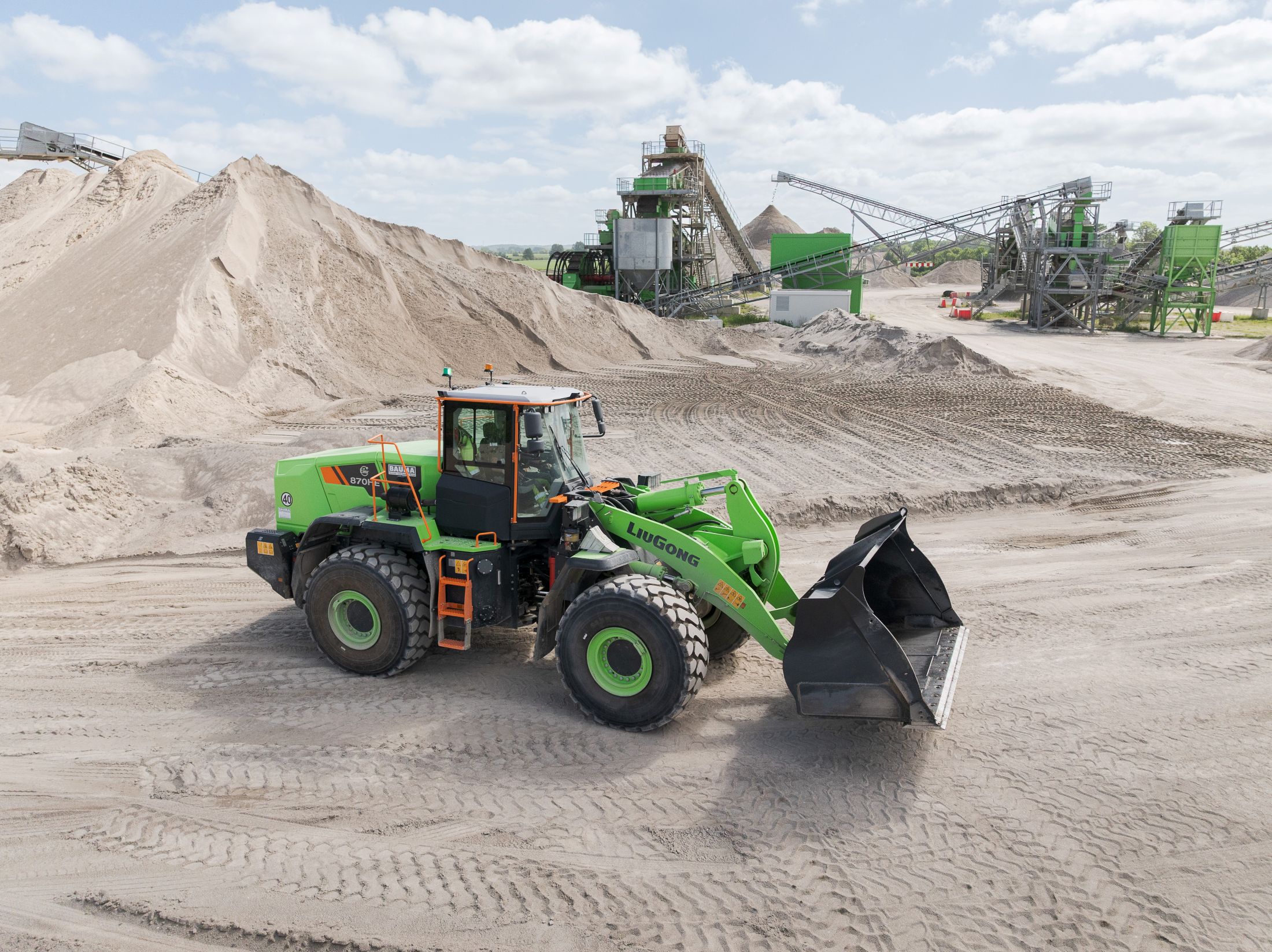 Ein grüner Radlader arbeitet auf einem Sand- und Kiesplatz vor einer industriellen Anlage.