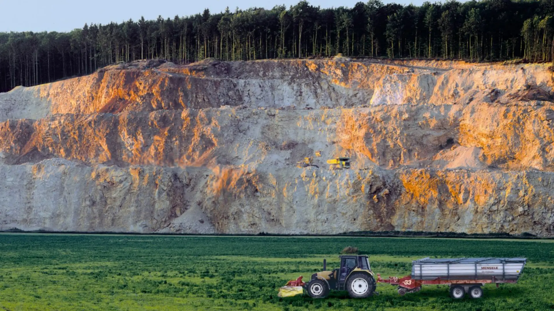 Ein Traktor mit einem Anhänger steht auf einer grünen Wiese im Vordergrund, während im Hintergrund eine große, steil abfallende Kalksteinformation zu sehen ist. Oben auf der Formation befindet sich ein dichter Wald. Die Abendsonne beleuchtet die Felswand teils orange-golden.