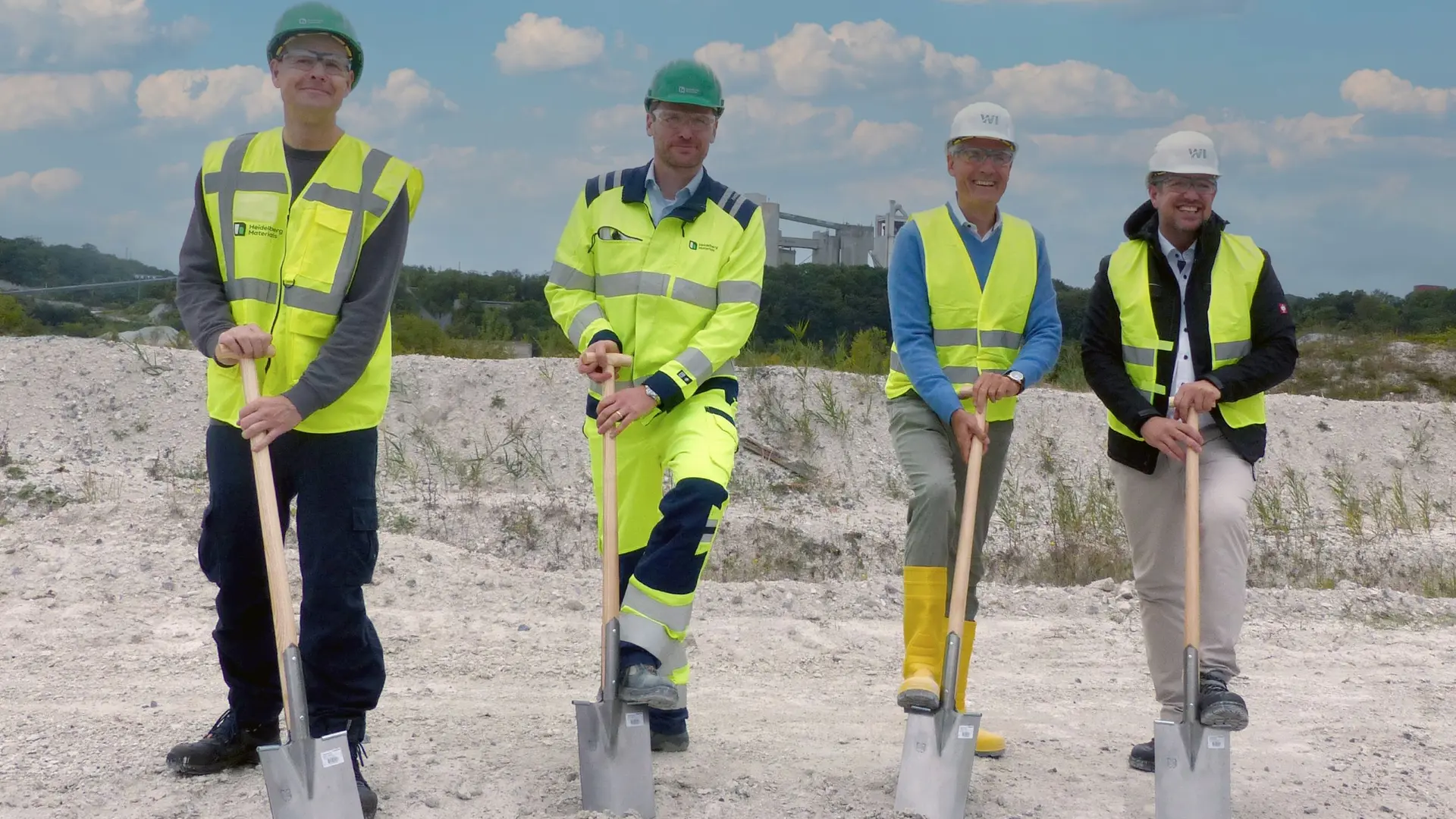 Vier Personen mit Schutzkleidung halten Spaten auf einer Baustelle.