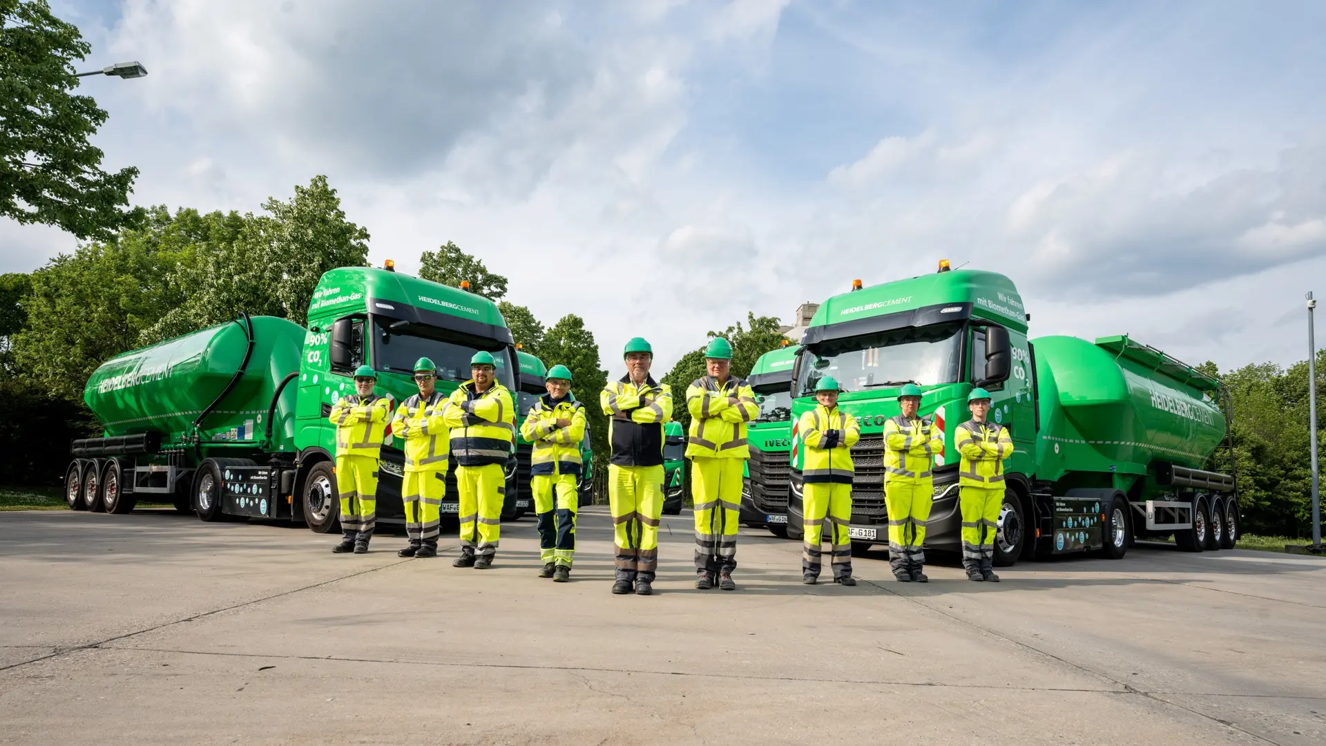 Gruppe von neun Personen in gelber Arbeitskleidung und Schutzhelmen vor zwei grünen Silozügen. Im Hintergrund sind Bäume und ein bewölkter Himmel sichtbar.