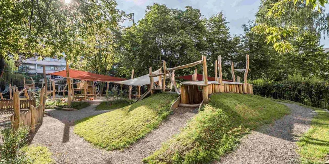 Spielplatz mit Holzstrukturen, Kieswegen und begrünten Erdhügeln, umgeben von Bäumen.