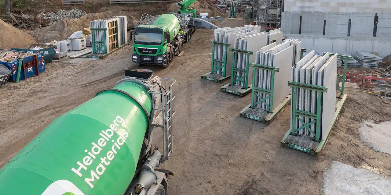 Grüne Betonmischer der Marke Heidelberg Materials auf einer Baustelle, umgeben von Betonfertigteilen auf Paletten; im Hintergrund sind Baugeräte, ein Kran und ein teilweise errichtetes Gebäude zu sehen.