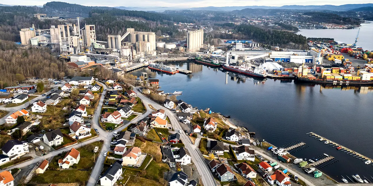 Luftaufnahme von Brevik in Norwegen mit dicht bebauten Wohnhäusern im Vordergrund, großem Zementwerk auf der linken Seite sowie einem Containerhafen rechts am Wasser. Im Hintergrund bewaldete Hügel und ein See.