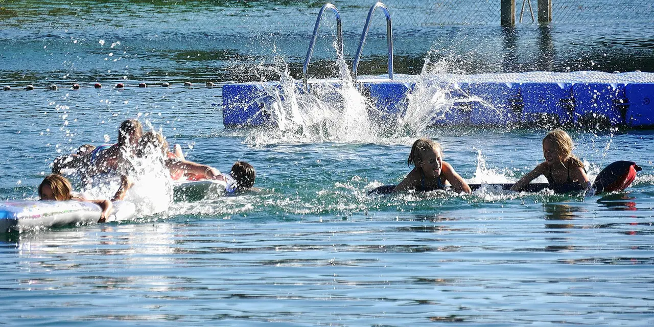 Kinder schwimmen in einem See. Im Hintergrund befinden sich ein Steg, eine Leiter und Bojen, die den Schwimmbereich abgrenzen. Spritzendes Wasser zeigt aktive Bewegung.