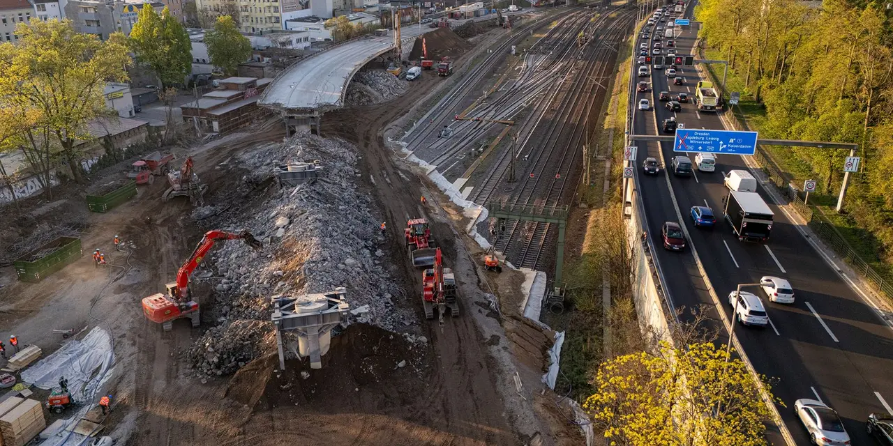 Luftaufnahme einer Baustelle neben einer Autobahn über Eisenbahnschienen in einer Stadt. Im linken Bereich ist ein großer Erdhügel mit Baggern und Bauarbeitern zu sehen. Rechts verlaufen dicht befahrene Fahrspuren der Autobahn parallel zu Gleisen. Im Hintergrund sind Stadtgebäude und Bäume im Abendlicht erkennbar.