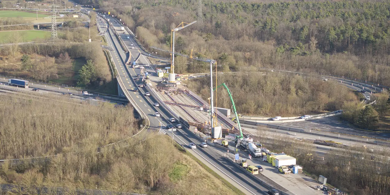 Luftaufnahme einer Autobahnbaustelle: Ein mehrspuriger Autobahnabschnitt mit laufenden Brückenarbeiten in der Bildmitte. Zwei große Kräne stehen auf der Baustelle, umgeben von Fahrzeugen und Baumaschinen. Verkehr fließt rechts und links um die Baustelle herum. Im Hintergrund sind Wälder, landwirtschaftliche Flächen und eine Stadt zu sehen.