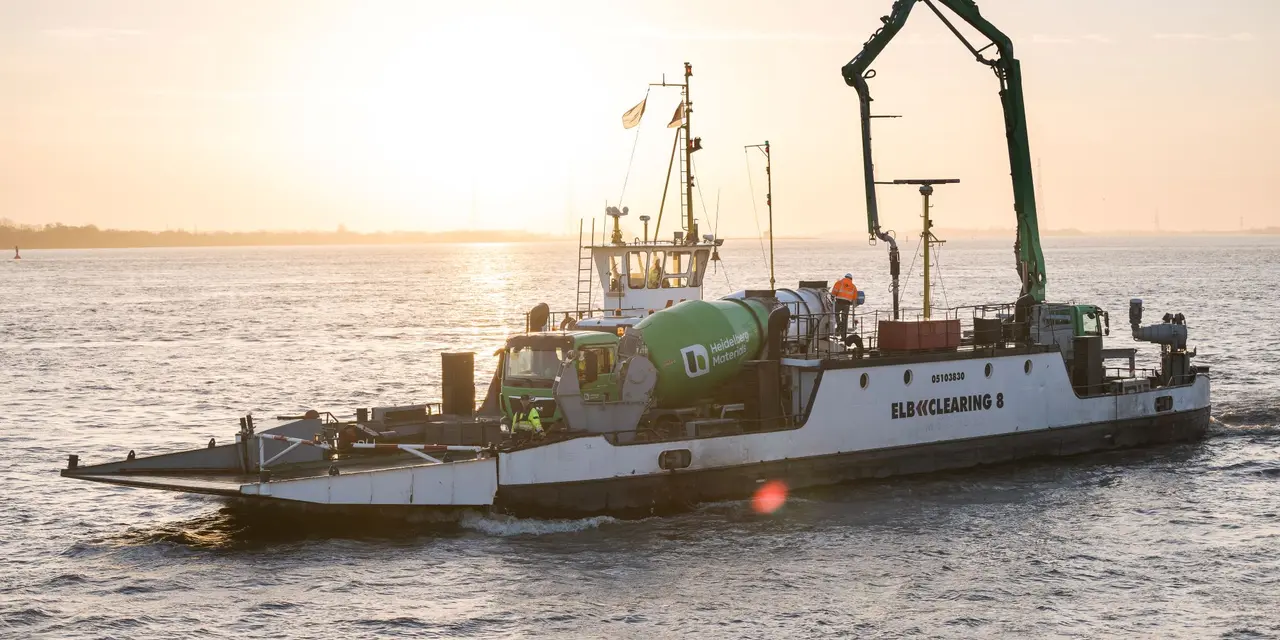 Fähre mit Betonmischer auf Deck fährt über das Wasser bei Sonnenuntergang, das Wasser reflektiert das Licht; im Hintergrund schwache Konturen einer Uferlinie sichtbar.