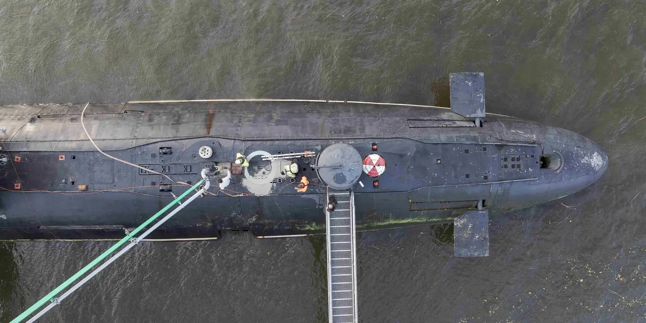Luftaufnahme eines U-Boots, das seitlich an einem Steg im Wasser liegt. Zwei grüne Schläuche verlaufen von der Plattform zum U-Boot, dessen Oberdeck sowie ein geöffneter Einstieg sichtbar sind. Vier Personen stehen um den Einstieg herum. Es wird betoniert.