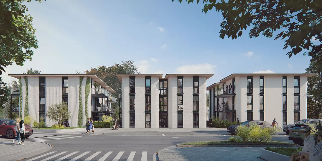 Moderne Wohnanlage mit drei weißen, mehrstöckigen Gebäuden, großen Fensterfronten und einigen vertikalen Begrünungen. Im Vordergrund ein Zebrastreifen, geparkte Autos und Gehwege, umgeben von Bäumen und Grünflächen. Blauer Himmel im Hintergrund.