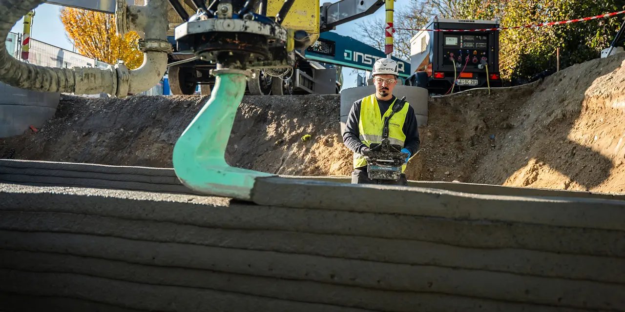 Ein Bauarbeiter überwacht einen 3D-Drucker, der Schichten aus Beton aufträgt, um einen Keller zu bauen. Im Hintergrund sind Baustellenfahrzeuge und herbstlich gefärbte Bäume sichtbar.