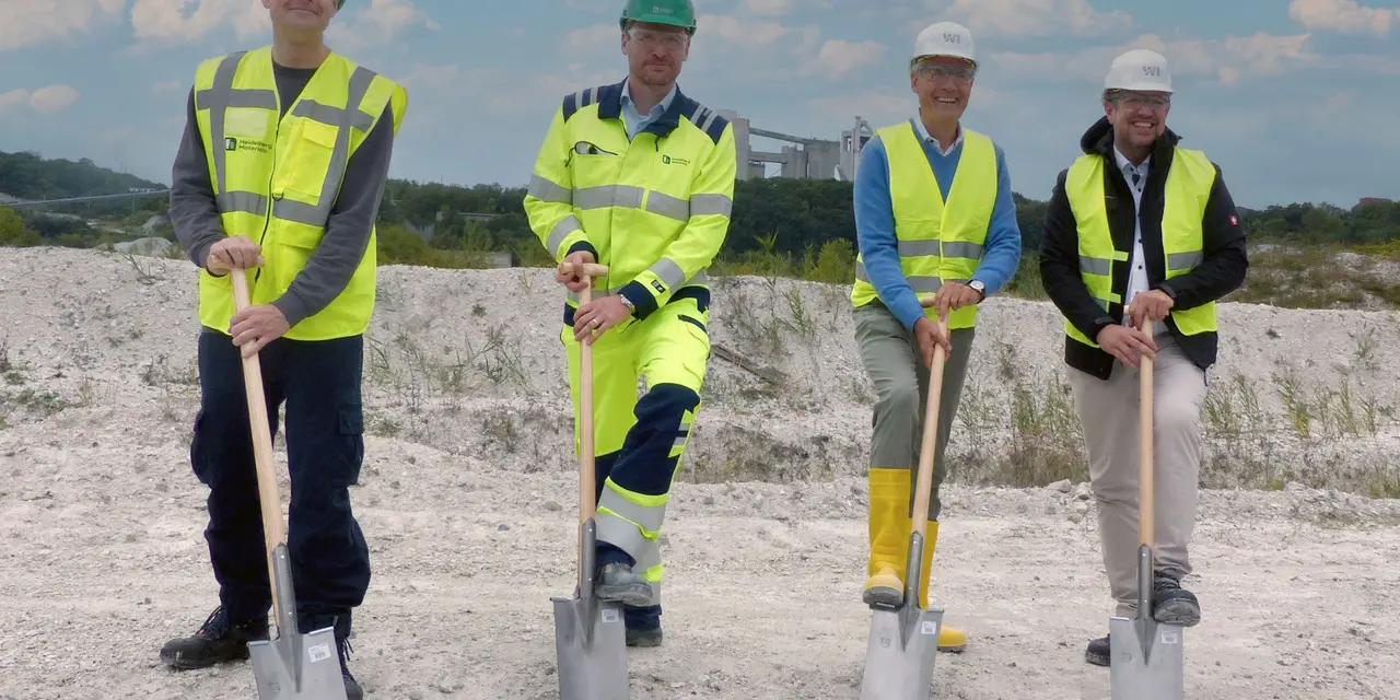 Vier Personen mit Schutzkleidung halten Spaten auf einer Baustelle.