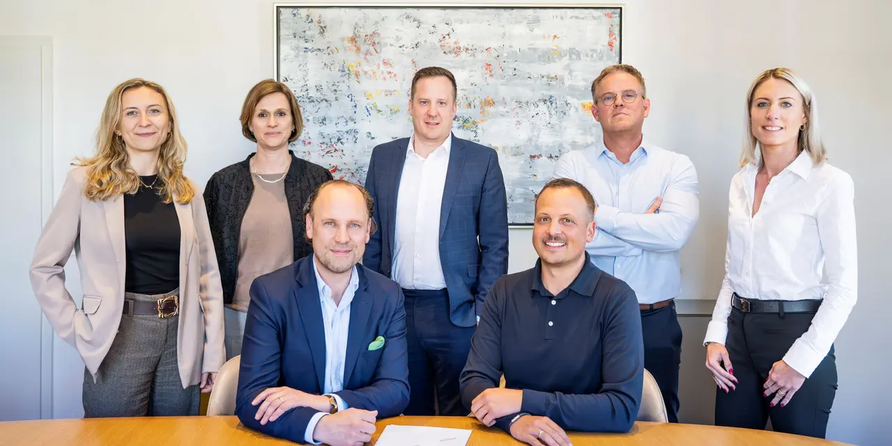 Mehrere Personen in Businesskleidung stehen und sitzen an einem Konferenztisch in einem Büro.