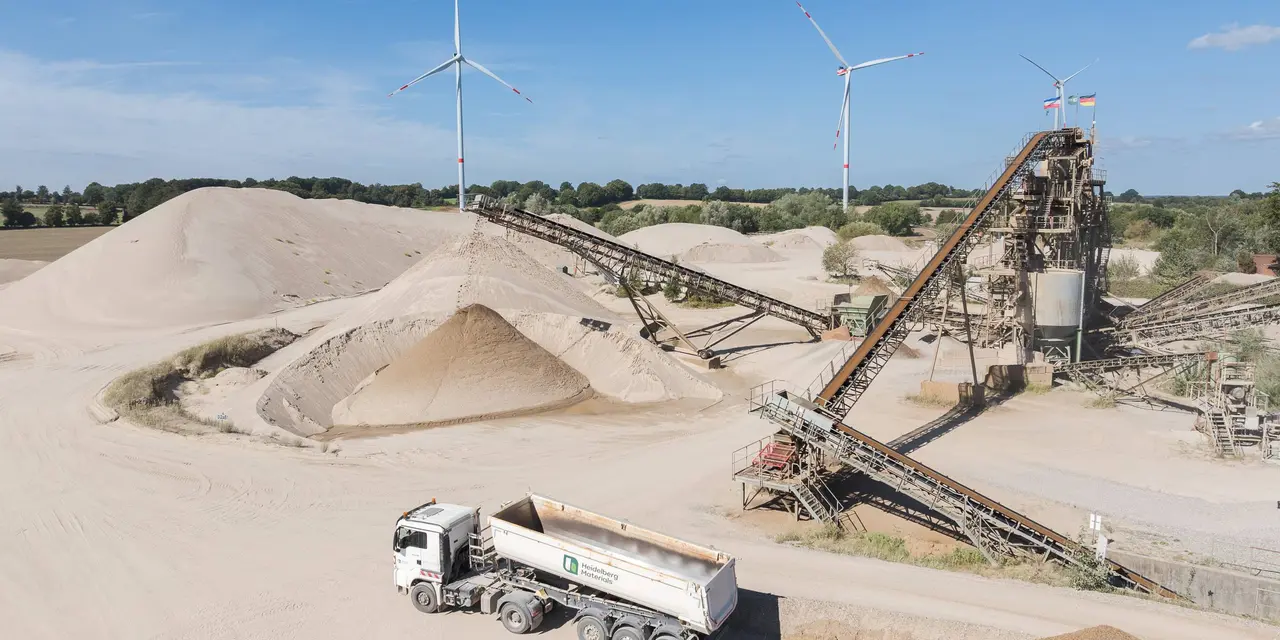 Ein Industriegelände mit Kieshaufen, Förderbändern und einem Lastwagen, im Hintergrund Windräder.