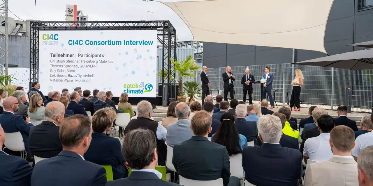 Gruppenveranstaltung im Freien mit einer Bühne, auf der mehrere Personen eine Präsentation vor einem sitzenden Publikum halten.