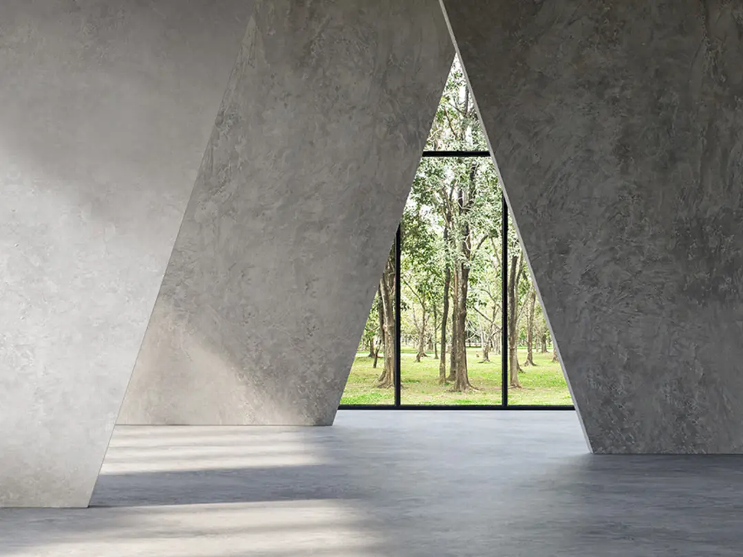 Ein moderner Innenraum mit geometrischen Betonwänden und großen Glasfenstern, durch die grüne Bäume sichtbar sind.