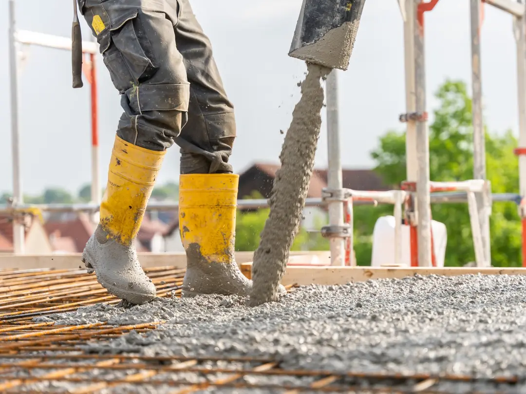 Arbeiter mit gelben Gummistiefeln gießt flüssigen Beton über ein Stahlarmierungsnetz auf einer Baustelle im Freien; Hintergrund zeigt Gerüst und unscharfe Landschaft.