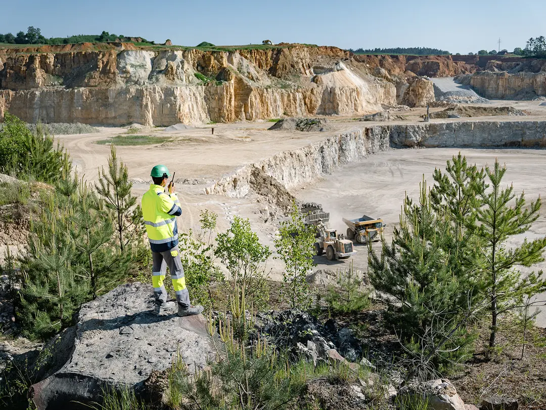 Eine Person in Sicherheitskleidung und Helm steht am Rand eines Steinbruchs von Heidelberg Materials. Im Hintergrund sind mehrere Fahrzeuge und gestufte Steinwände zu sehen. Zu beiden Seiten sind Büsche und Bäume, die den natürlichen Rand der Umgebung betonen.