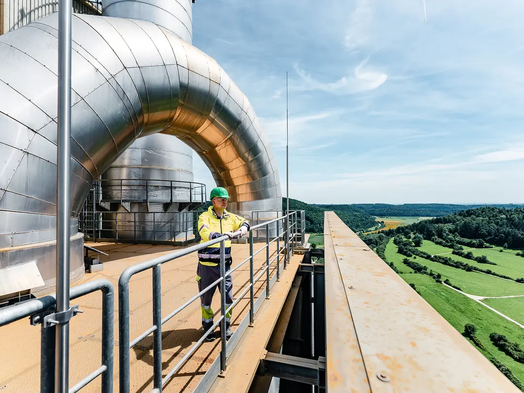 Ein Arbeiter in Schutzkleidung und Helm steht auf einer erhöhten Plattform im Zementwerk. Im Hintergrund sind eine grüne Landschaft mit Feldern und Wald sowie ein blauer Himmel sichtbar.
