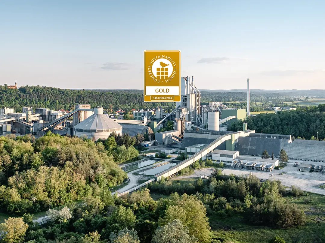Luftaufnahme eines des Zementwerks Burglengenfeld inmitten einer grünen Landschaft mit Wald und Wiesen. Zentral über dem Werk wird das CSC Zertifikat gezeigt.