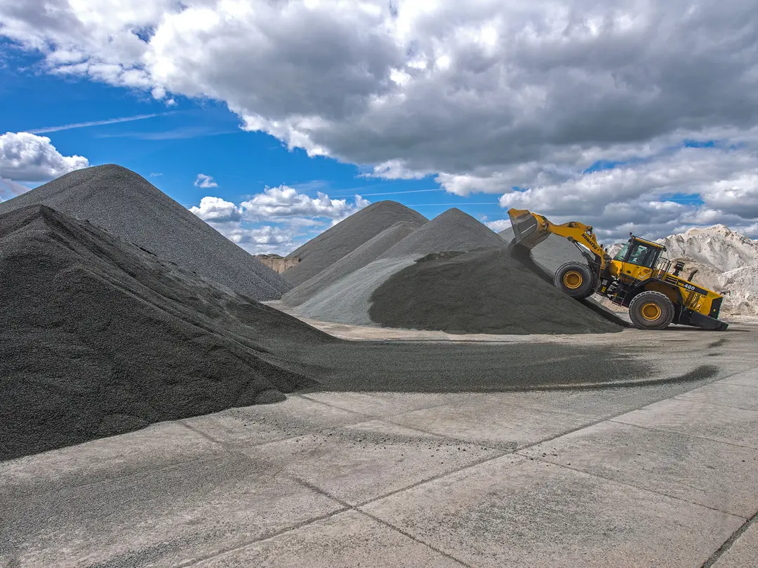 Gelber Radlader an einer grauen Kieshalde im Kieswerk, umgeben von mehreren Kieshaufen; Himmel bedeckt mit Wolken.