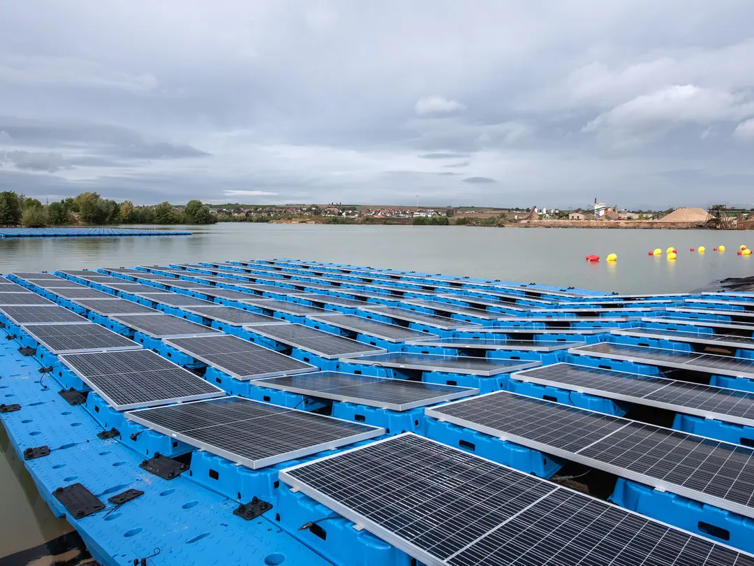 Schwimmende Solaranlage auf einem Baggersee, bestehend aus blauen Plattformen mit montierten Solarmodulen. Im Hintergrund sind Bäume, Industriebauten und gelbe Bojen sichtbar. Der Himmel ist bewölkt.