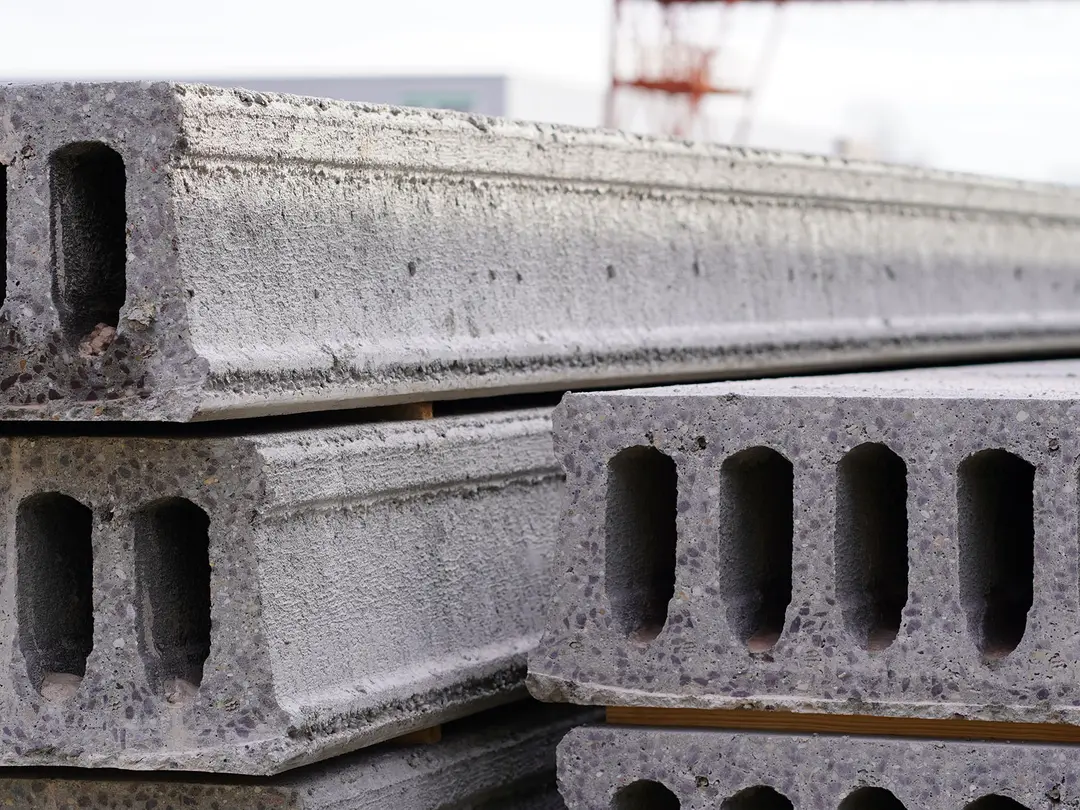 Nahaufnahme von vorgefertigten Spannbetonhohldecken mit rechteckigen Hohlräumen, die für Bauprojekte verwendet werden; im Hintergrund unscharf ein Baukran erkennbar.