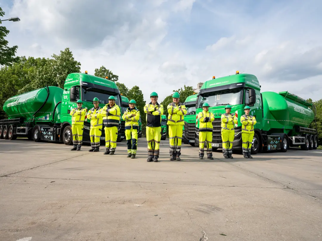 Gruppe von neun Personen in gelber Arbeitskleidung und Schutzhelmen vor zwei grünen Silozügen. Im Hintergrund sind Bäume und ein bewölkter Himmel sichtbar.