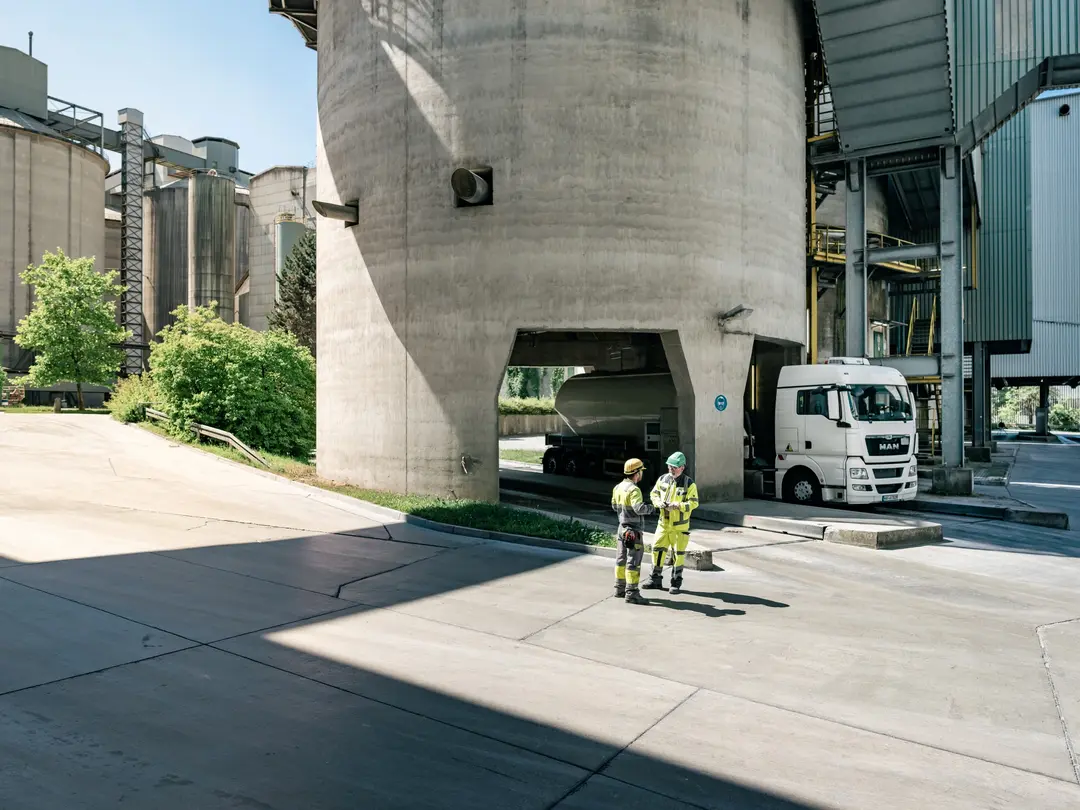 Zwei Arbeiter in Schutzkleidung stehen auf einem Zementwerk und unterhalten sich; im Hintergrund ein weißer Silozug.