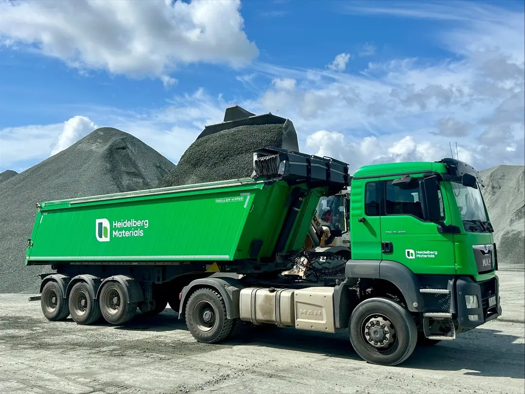 Ein grüner Lastwagen von Heidelberg Materials steht vor einem Kies- oder Schotterhaufen auf einem Industriegelände.