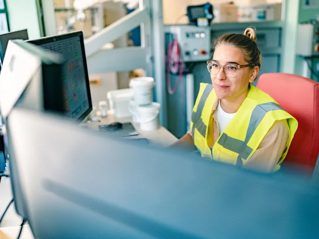 Mitarbeiterin in Sicherheitsweste arbeitet am Computer in einem industriellen Büro.