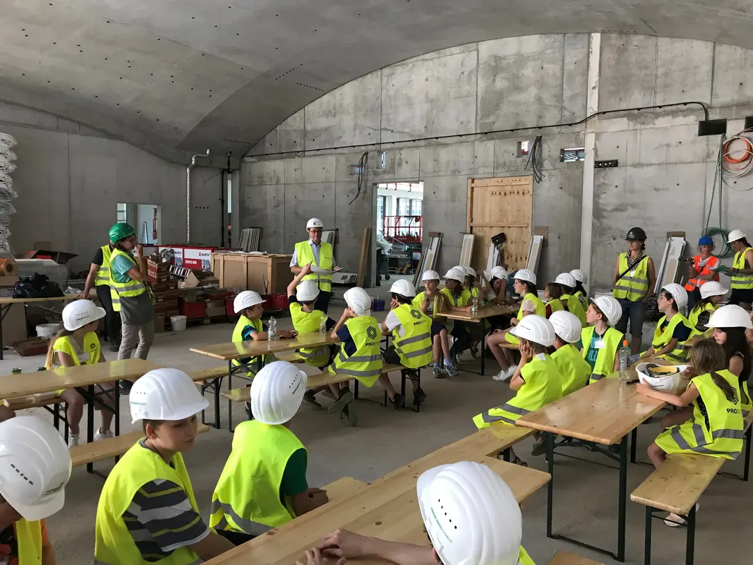 Kinder mit Schutzhelmen und Warnwesten hören in einer Baustellenumgebung einer Präsentation zu.