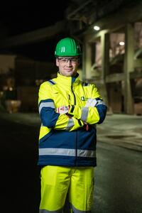 Ein Mitarbeiter in Sicherheitskleidung und grünem Schutzhelm steht vor einem Industriegebäude bei Nacht.