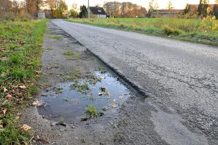 Schmale Landstraße mit Schlaglöchern und einer Wasserpfütze am Rand in ländlicher Umgebung.