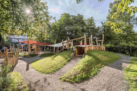 Spielplatz mit Holzstrukturen, Kieswegen und begrünten Erdhügeln, umgeben von Bäumen.