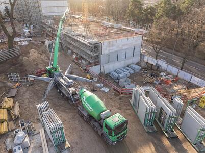 Baustelle mit halb fertiggestelltem Gebäude im Hintergrund, Betonmischer und Betonpumpe von Heidelberg Materials im Vordergrund, sowie gestapelte Wandmodule rechts. Baumaterialien und Baugeräte links und unten verteilt. Baustelle von oben bei Tageslicht mit umliegenden Bäumen.